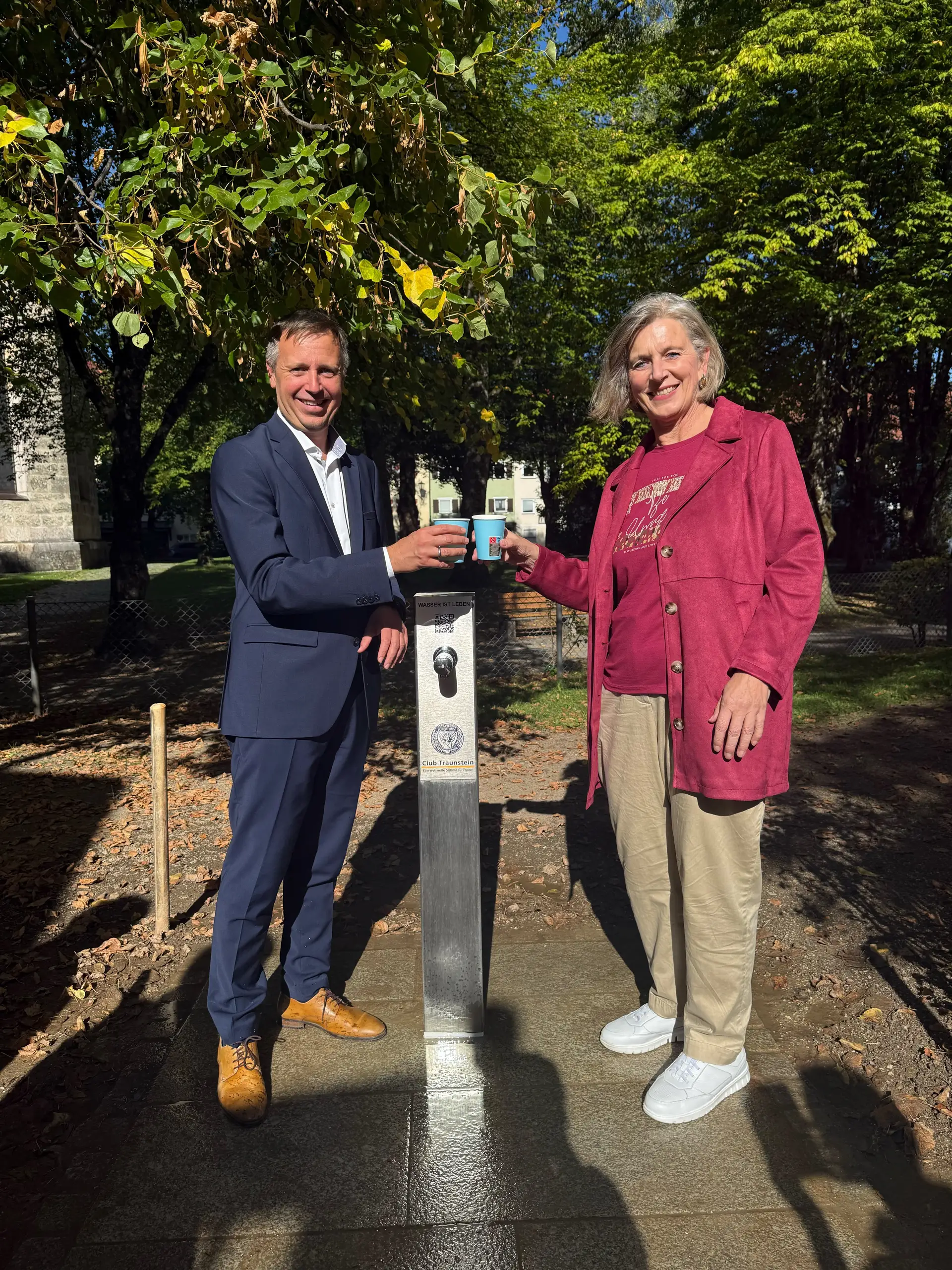 Zwei Personen stoßen an einem öffentlichen Trinkwasserbrunnen am Spielplatz in der Au auf die Spende des Soroptimist International Club Traunstein an.